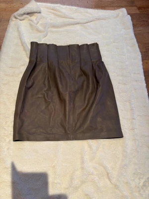 Brun skinnkjol från Saint Tropez - Snygg brun kort skinnkjol från Saint Tropez i storlek M. Kjolen har hög midja med breda veck upptill och en diskret dragkedja bak. Materialet är mjukt och ger en cool vibe, perfekt för dig som gillar stilrena plagg med edge. Färg: mullvad. Skick : 10/10