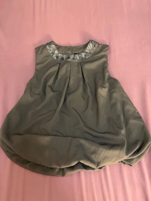 Grå blus med paljetter vid halsen - Säljer en snygg grå blus med rundad hals som är dekorerad med silvriga paljetter. Blusen är ärmlös och har en bubblig, voluminös form nedtill. Stängs med två knappar i nacken. Perfekt för dig som gillar lite extra bling på toppen! SER SNYGGARE UT PÅ!!💕💕