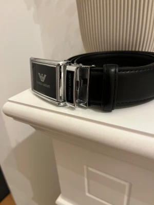 Svart skärp från Giorgio Armani - Snyggt svart bälte från Giorgio Armani med stilren silverfärgad spänne och logotyp på framsidan. Bältet är tillverkat i skinn och har en klassisk, slät design som ger en exklusiv känsla. Perfekt för dig som vill ha en clean och lyxig accessoar.