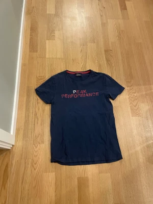 Mörkblå Peak Performance t-shirt - Snygg mörkblå t-shirt från Peak Performance med röd text på bröstet och en röd detalj på insidan av kragen. Klassisk rund hals och korta ärmar. Perfekt för dig som gillar sportiga och stilrena plagg.