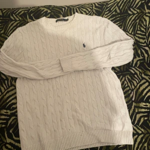 Vit stickad tröja från Polo Ralph Lauren - Säljer en vit stickad tröja från Polo Ralph Lauren med klassiskt kabelstickat mönster och rund hals. Tröjan har långa ärmar och den ikoniska blå Polo-loggan broderad på bröstet. Perfekt för dig som gillar stilren och tidlös design.