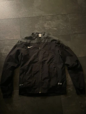 Svart Nike vindjacka Dri-FIT - Säljer en svart Nike vindjacka med Dri-FIT-tyg och dragkedja framtill. Jackan har hög krage, två fickor med dragkedja och klassisk Nike Swoosh på bröstet. Perfekt för dig som vill ha en clean och sportig look. Den är i fint skick och har ett fantastiskt pris. Kontakta vid frågor!