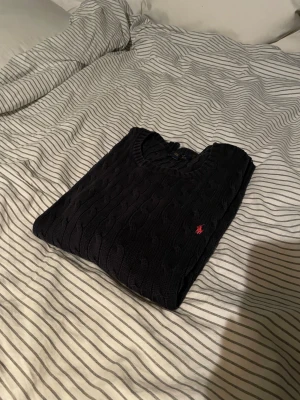 Marinblå stickad tröja Polo Ralph Lauren - Snygg marinblå stickad tröja från Polo Ralph Lauren i storlek XL. Tröjan har kabelstickat mönster, rund hals och långärmade ribbade muddar. På bröstet finns den klassiska röda Polo-loggan broderad. Perfekt för dig som gillar stilrena och tidlösa plagg.
