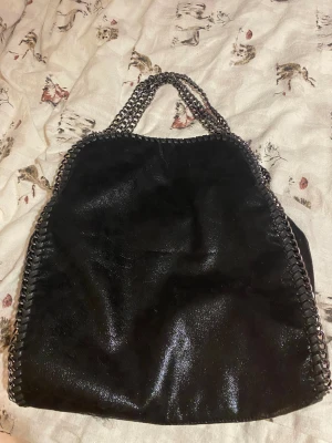 Svart axelväska med kedjehandtag, Liknar en Stella Mccartney väska - Snygg stockholmsstil svart axelväska med glansig yta och grova kedjehandtag i metall. Ser exakt ut som en Stella Mccartney väska. Vet ej vart den är köpt från, fick den av min syster. Väskan har dekorativa kedjor längs kanterna och ett rymligt innerfack med dragkedja. Den är helt ny, aldrig använd. Säljes då den inte kommer till användning ❤️Hör av er ifall ni har frågor☺️