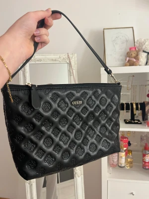 Svart quiltad axelväska från Guess - Snygg svart axelväska från Guess med quiltad yta och präglade loggor över hela väskan. Väskan har guldfärgade detaljer, dragkedja upptill och ett ljust innerfoder med Guess-tryck. Perfekt storlek för det viktigaste och enkel att bära över axeln.