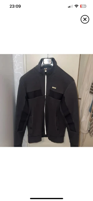 Boss hoodie/kofta - Svart zipjacka från BOSS med stilren design och hög krage. Jackan har vita detaljer vid dragkedjan och BOSS-logga på bröstet. Materialet är mjukt och känns sportigt, perfekt för chill dagar. Långärmad och med fickor på sidorna, nypris 2100kr, boss loggan blev lite sliten i tvätten så jag har limmat fast loggan så den sitter bra men annars är den väldigt fin