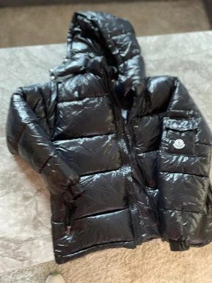 Svart Moncler Maya - Säljer en svart pufferjacka från Moncler med glansig finish och huva. Jackan har dragkedja framtill, ficka med Moncler-logga på ärmen och quiltad design. Perfekt för dig som vill ha en varm och snygg jacka med streetkänsla.