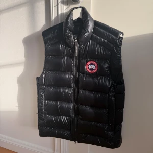 Svart dunväst från Canada Goose - Snygg svart dunväst från Canada Goose med klassisk logga på bröstet. Västen har hög krage, quiltad design och dragkedja framtill. Perfekt för lager-på-lager och håller dig varm under kyliga dagar. Stilsäker och ikonisk look.