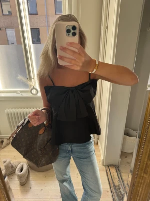 Svart linnetopp med stor rosett - Säljer en svart linnetopp med smala axelband och en stor rosett framtill. Toppen har en lätt utsvängd form och är ärmlös, vilket ger en snygg och trendig siluett. Perfekt att styla med jeans eller kjol för en cool look.