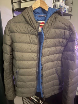 Grå Colmar pufferjacka med huva - Säljer en grå pufferjacka från Colmar med huva och blått foder. Jackan har dragkedja framtill, två sidofickor med dragkedja och Colmar-logga på ärmen. Perfekt för dig som vill ha en clean och sportig look under höst och vinter. OBS SISTA BILDEN. Ser värre ut än vad det är.
