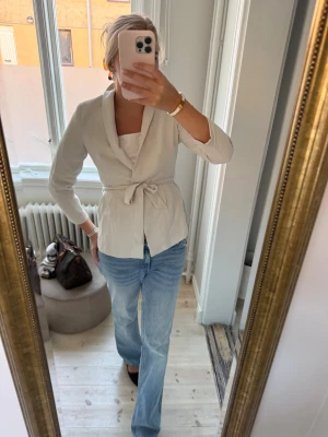 Beige kavaj med knytband - Snygg beige kavaj med knytband i midjan och klassisk krage. Jackan har lång ärm och en stilren, feminin siluett. Perfekt att styla med jeans eller kjol för en clean och modern look. Liten i storlek 