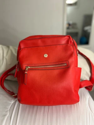 Röd Don Donna ryggsäck - Snygg röd Don Donna ryggsäck i skinnimitation med flera fack och guldiga dragkedjor. Insidan är vit med röda prickar, vilket ger en cool kontrast. Justerbara axelremmar och praktiska sidofickor gör den perfekt för dig som vill ha både stil och funktion.