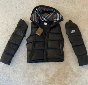 Svart pufferjacka från Burberry M - Svart pufferjacka från Burberry med avtagbara ärmar och huva. Insidan har klassiskt rutigt Burberry-foder i grått, vitt och rött. Jackan har dragkedja, knappar och två sidofickor. Stor Burberry London England-patch på ena ärmen.