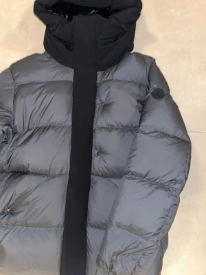 Svart Moncler pufferjacka med huva - Säljer en stilren svart pufferjacka från Moncler med stor huva och diskret logga på ärmen. Jackan har breda quiltade sektioner, dragkedja och tryckknappar framtill. Perfekt för kalla dagar och har en loose passform som ger en chill vibe.