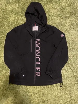 Svart vindjacka från Moncler - Svart vindjacka från Moncler med stor logga i rosa längs dragkedjan. Jackan har huva, tre framfickor med dragkedja och Moncler-märke på ärmen. Snygg och stilren design med elastiska muddar och justerbar nederkant.
