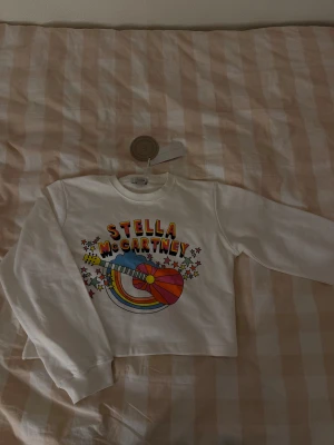 Vit sweatshirt Stella McCartney Kids - Cool vit sweatshirt från Stella McCartney Kids med färgglatt tryck av gitarr, regnbåge och stjärnor på bröstet. Rund hals och långa ärmar. Tillverkad i mjuk bomull med hållbarhetsmärkning. Perfekt för dig som gillar färg och statement-plagg. Helt oanvänd med prislapp kvar.