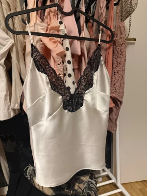 Vit satin linnetopp med spets - Säljer en snygg vit linnetopp i satin med tunna axelband och djup v-ringning dekorerad med svart spets. Toppen har en glansig finish och sitter lätt och luftigt på kroppen. Perfekt för dig som gillar en chic och feminin stil.