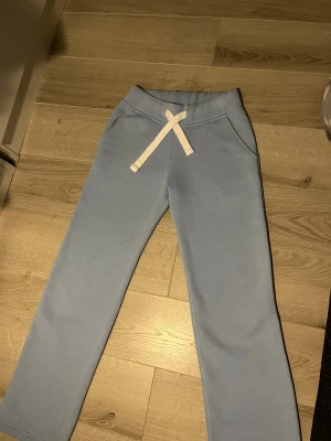 Ljusblå mjukisbyxor från Russedress - Ljusblå mjukisbyxor från Russedress med vita snören i midjan och bekväma fickor. Byxorna är raka i modellen och tillverkade i mjukt bomullsmaterial, perfekta för chill dagar. Snygg och enkel design som passar till hoodie eller t-shirt.