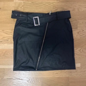 Svart skinnkjol med bälte och dragkedja - Snygg svart kortkjol i skinnimitation med bälte och stor silverfärgad spänne