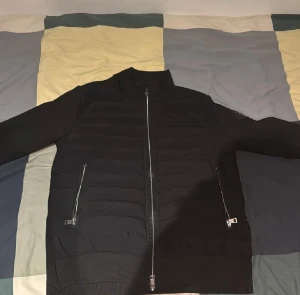 Svart Moncler pufferjacka herr S - Snygg svart pufferjacka från Moncler Grenoble med quiltad front, två dragkedjefickor och Moncler-logga på ärmen. Jackan har hög krage och hel dragkedja framtill. Materialmix med polyester och stickade detaljer ger en modern look.