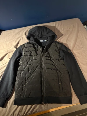 Svart cardigan från Polo Ralph Lauren - Svart pufferjacka från Polo Ralph Lauren med huva och snyggt quiltad front. Jackan har dragkedja, två fickor med knappstängning och ett Polo-märke på ärmen. Ärmar och huva är i mjuk bomull, medan framsidan är i glansig syntet. Perfekt för höst och vinter.