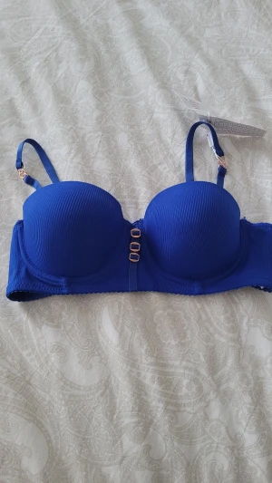 Blå balconette behå från Dandissi - Snygg blå behå från Dandissi med lätt vadderade kupor och justerbara axelband. Framsidan har tre dekorativa metallspännen i guld och tyget är ribbat för extra stil. Perfekt för dig som vill ha en modern och bekväm look 