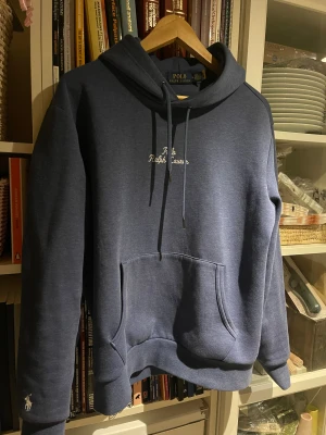 Blå hoodie från Polo Ralph Lauren - Snygg mörkblå hoodie från Polo Ralph Lauren med broderad logga på bröstet och klassisk snörning i huvan. Tröjan har en stor magficka och ett litet broderat Polo-märke vid ärmslutet. Perfekt för chill dagar och enkel att matcha med jeans.