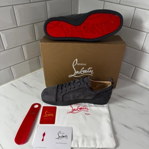 Grå mocka sneakers från Louboutin - ‼️GÅR INTE NER I PRIS‼️Snygga grå sneakers från Christian Louboutin i mjuk mocka med klassisk röd sula. Skorna har rund tå, platt sula och snörning framtill. Insidan är i beige skinn med guldfärgad logga. Perfekt för dig som gillar exklusiv och stilren design.