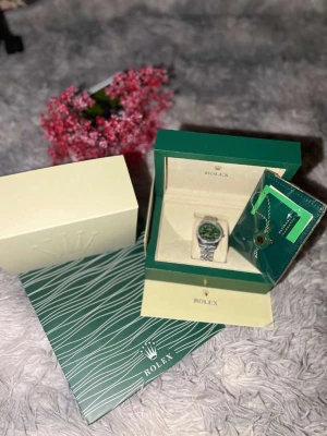 Rolle atejust med grön urtavla - Exklusiv Rolle Datejust med grön urtavla och silverfärgad länk i metall. Klockan har en rund form, glittrande stenar runt boetten och klassisk design. Levereras i originalbox med certifikat och tillbehör. Perfekt för dig som gillar lyxiga accessoarer.