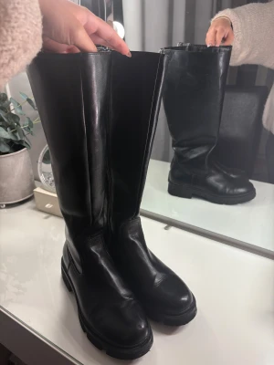 Svarta höga stövlar med dragkedja - Säljer ett par svarta höga stövlar i skinn med grov platt sula och rund tå. Stövlarna har dragkedja baktill för enkel på- och avtagning. Stilrena och chunky, perfekta för att lyfta din outfit under höst och vinter.