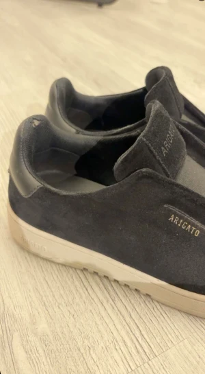 Svarta Arigato sneakers i mocka - Svarta sneakers från Arigato med stilren design och beige platt sula. Ovandelen är i mjuk mocka med diskret Arigato-logga i guld på sidan och plösen. Rund tå och elastisk panel för enkel påtagning. Perfekt för dig som gillar minimalistisk stil. 