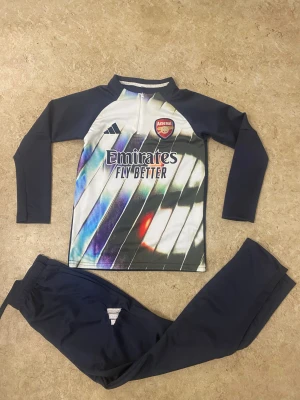 Arsenal träningsset från Adidas - Säljer ett Arsenal träningsset från Adidas med långärmad tröja och byxor. Tröjan har half zip, klubbmärke och coolt randigt mönster i vitt, blått och inslag av rött. Byxorna är marinblå med Adidas-logga. Perfekt för fotbollsträning eller chill.