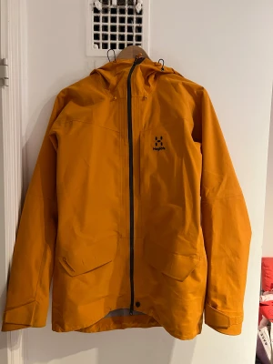 Haglöfs skidjacka - Säljer en snygg gul skidjacka från Haglöfs med huva och hel dragkedja framtill. Jackan har två stora fickor med lock, justerbara ärmslut och en clean design. Perfekt för dig som gillar outdoor och vill sticka ut med färg.