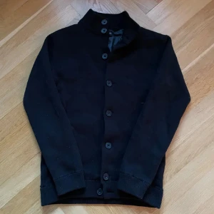 Svart cardigan  - Svart stickad cardigan med knappar hela vägen framtill. Tröjan har två fickor med ribbade detaljer och långa ärmar. Perfekt för dig som gillar enkel och stilren design med lite edge. Passar S/M
