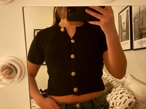 Svart croppad topp med guldknappar - Säljer en svart croppad topp med korta ärmar och snygga guldknappar framtill. Toppen har ett diskret kabelstickat mönster och ribbad kant nedtill. Från HM, säljes för 130kr 
