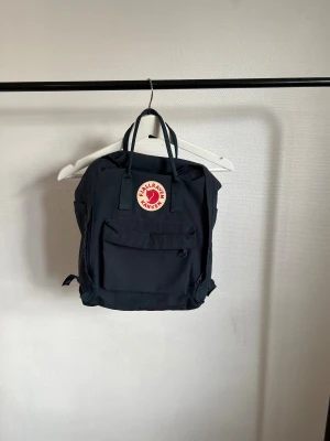 Mörkblå Fjällräven Kånken ryggsäck - Fjällräven Kånken ryggsäck i färgen mörkblå. Sittunderlaget finns kvar och är aldrig använd. Har använts men är i väldigt fint skick. Lite trådig på insidan men inget märkbart. Är i storlek 36 cm. Antar att den är äkta då jag fick den som present!