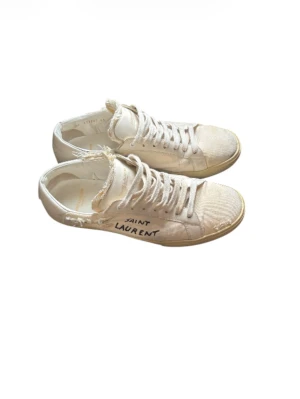Saint Laurent vita sneakers med rå kant - Säljer ett par vita sneakers från Saint Laurent med cool rå kant och handskriven logga på sidan. Skorna har snörning, platt sula och rund tå. Materialet är canvas och skinn, vilket ger en schysst kontrast. Perfekt för dig som gillar en edgy och avslappnad stil.