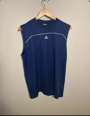 Marinblå ärmlös linnetopp från Adidas - Säljer en marinblå ärmlös linnetopp från Adidas med vit piping och logga på bröstet. Toppen har rund hals och är tillverkad i mjuk polyester, perfekt för träning eller sport. Enkel och stilren design som ger en sportig vibe.