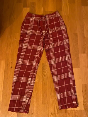 Röd-rutiga byxor från Kappahl XS - Snygga röd-rutiga byxor från Kappahl i storlek XS. De har ett klassiskt rutmönster i vitt och rött, elastisk midja med snörning och raka ben. Perfekta för chill dagar i Jul eller när du vill ha en avslappnad look. 