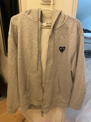 Grå hoodie Comme des Garçons Play - Grå hoodie från Comme des Garçons Play med dragkedja, huva och snörning. Ikoniskt svart hjärta med ögon på bröstet och stort tryck på ryggen. Två fickor fram och ribbade muddar. Perfekt för dig som gillar streetwear och vill sticka ut.