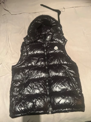 Svart dunväst från Moncler med huva - Snygg svart dunväst från Moncler med glansig finish och avtagbar huva. Pris är ej hugget i sten vid snabb och smidig affär 