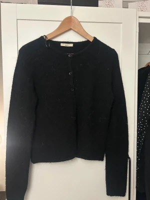 Svart fluffig kofta från Gina Tricot - Svart kofta från Gina Tricot i storlek S. Koftan har rund hals, knappar framtill och är tillverkad i ett fluffigt stickat material.