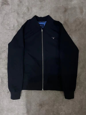 Svart bomberjacka från GANT - Storlek 11-12 år. Mörkblå junior bomberjacka från GANT med stilren design och silverfärgad dragkedja framtill. Jackan har ribbade muddar och en ljusblå insida med märkning. Perfekt för era barn.