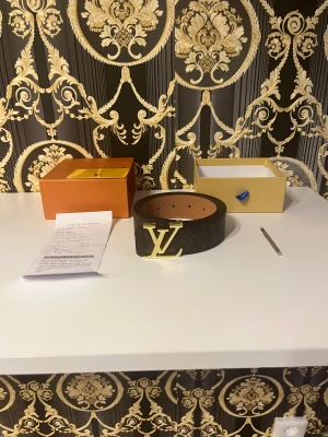 Louis Vuitton mörkbrunt skärp med LV-spänne - Exklusivt mörkbrunt Louis Vuitton-bälte i äkta skinn med ikoniskt guldigt LV-spänne. Bältet har klassiskt monogrammönster och är tillverkat i Frankrike. Perfekt accessoar för dig som vill sticka ut med lyxig stil. Kommer med originalask och kvitto. OBS PRIS KAN DISKUTERAS SNABB AFÄRR