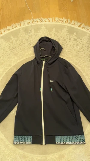 Svart hoodie från BOSS med dragkedja - Svart hoodie från BOSS med vit dragkedja och huva. Långärmad modell med snörning i huvan och BOSS-logga på bröstet. Nederkanten har ett coolt mönster med upprepade BOSS-tryck i turkos och vitt. Perfekt för chill dagar.