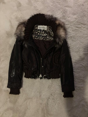 Brun bomberjacka med fuskpäls från Alexandra - Säljer en mörkbrun bomberjacka från Alexandra med croppad passform, stickade muddar och stor krage i fuskpäls. Insidan har leopardmönstrat foder och jackan stängs med dragkedja. Snygga metallringar och detaljer vid midjan ger extra edge.