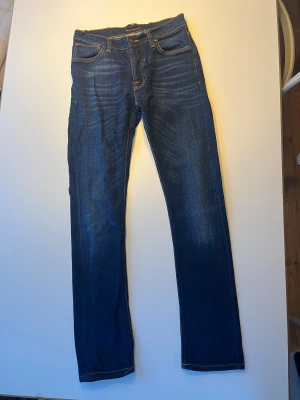 Mörkblå jeans från Nudie Jeans - Säljer ett par mörkblå jeans från Nudie Jeans med klassisk femficksdesign och kontrastsömmar. Modell Dude Dan med lite fabricerade slitningar. Mycket fint skick. 27/32 men lite större i storleken mer som 29/32