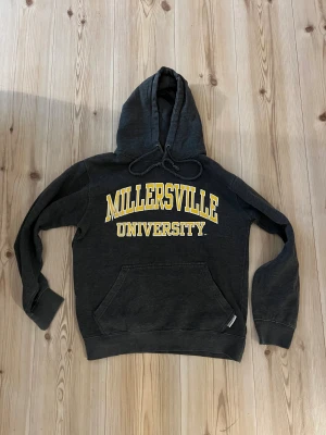 MSP EIGHTY8 grå hoodie - strl.S - Snygg mörkgrå hoodie från MSP Eighty3 med Millersville University-tryck i gult och vitt på bröstet. Tröjan har huva med snörning, stor magficka och ribbade muddar. Perfekt för dig som gillar college-vibe och vill ha en bekväm och stilren look.