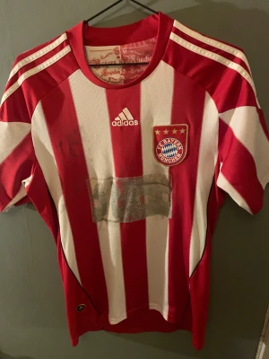 Bayern München rödvit Adidas fotbollströja - Säljer en officiell Bayern München fotbollströja från Adidas med klassiska röda och vita vertikala ränder, klubbmärke med fyra stjärnor på bröstet och tryck på ryggen. Tröjan har korta ärmar och är tillverkad i lätt, ventilerande polyester.