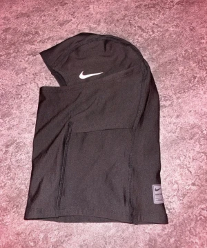 Svart Nike Pro Skimask  - Nike Pro balaclava i svart med vit logga framtill. Tillverkad i mjukt och stretchigt syntetmaterial som andas, perfekt för träning utomhus. Snygg och sportig design som täcker hela huvudet och halsen. Märke syns på etikett nedtill.